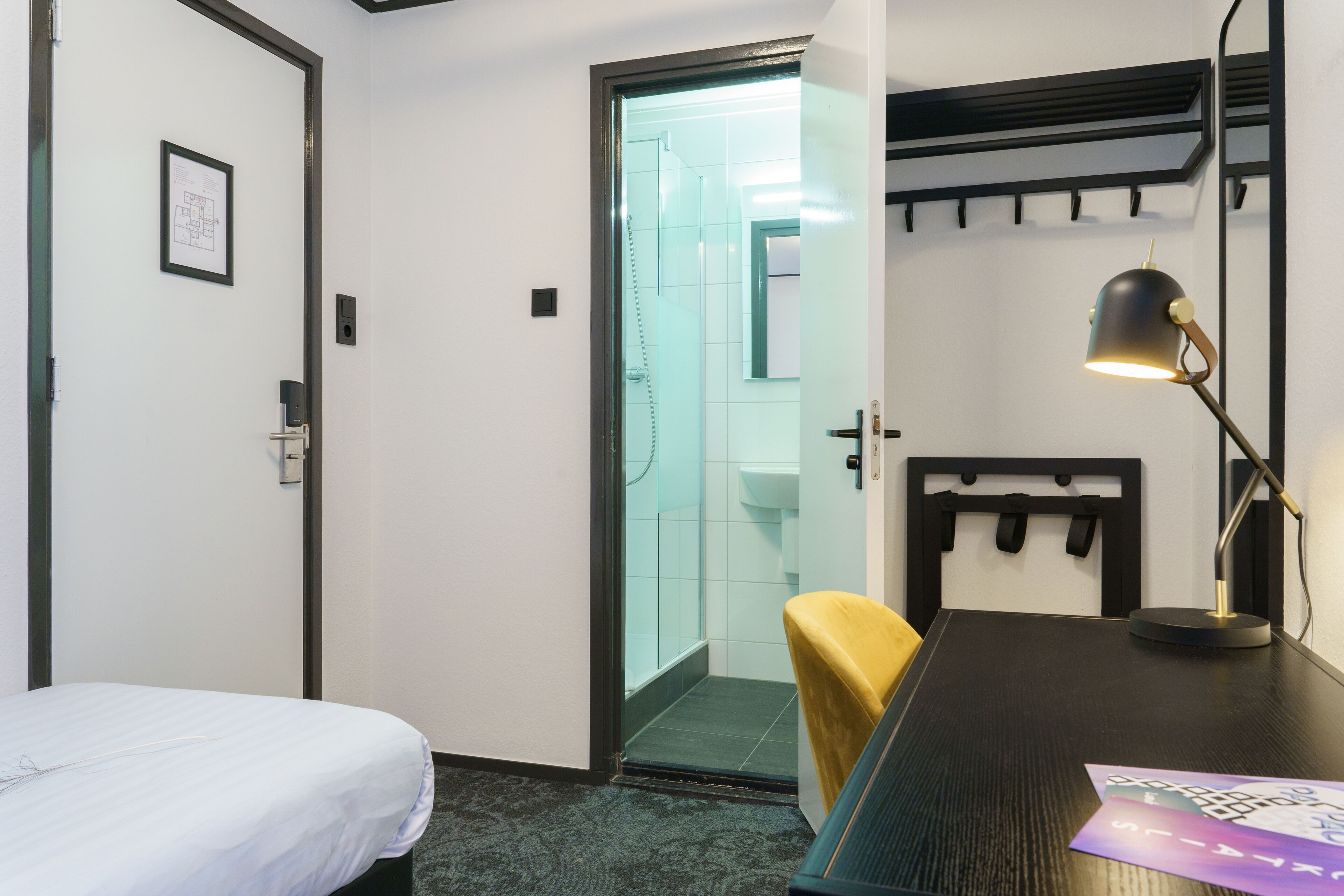 A hotel room with a bed, a desk, a chair, a desk lamp, and a bathroom visible through an open door.