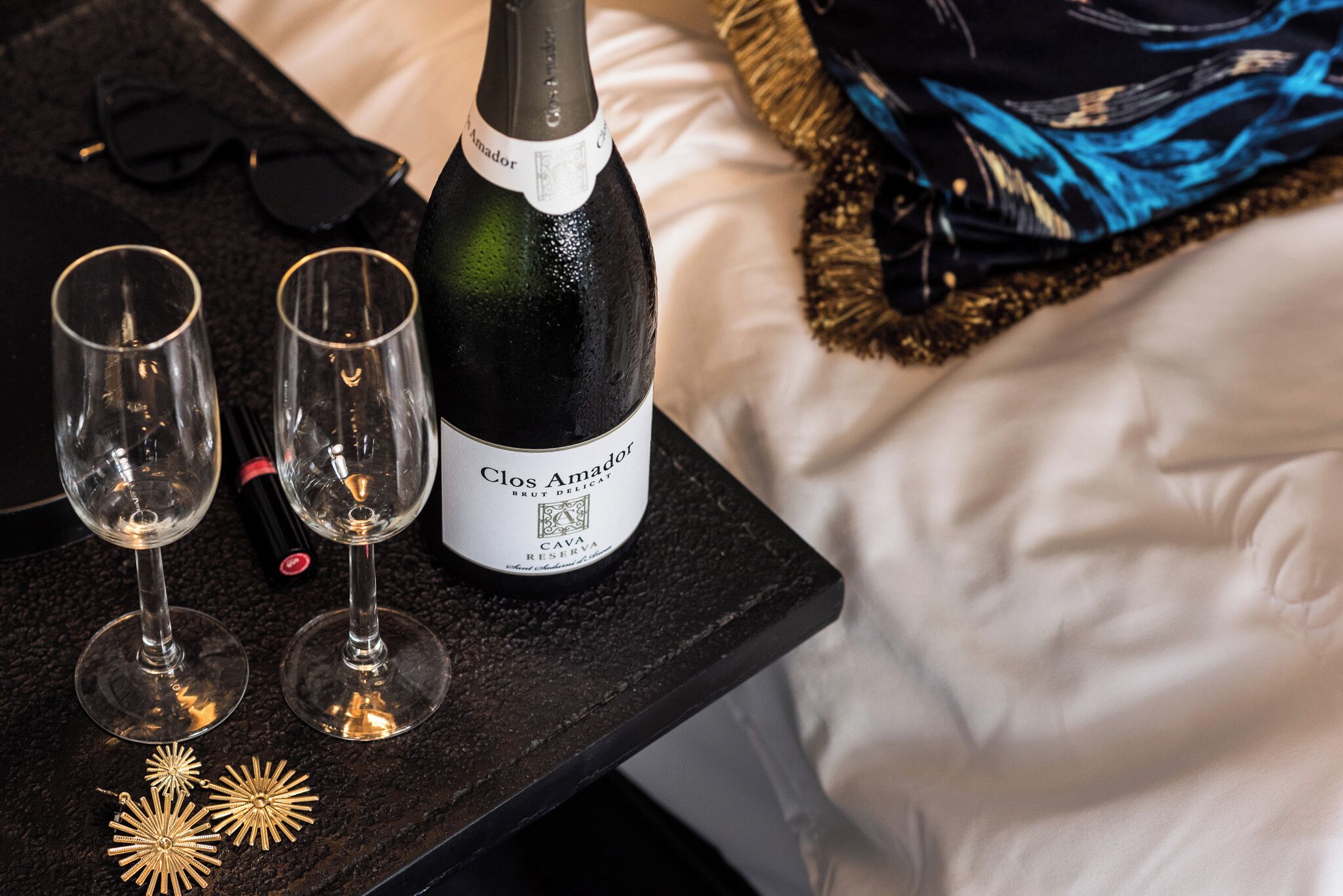 A bottle of Clos Amador Brut on a black table with two empty wine glasses and decorative golden flowers.