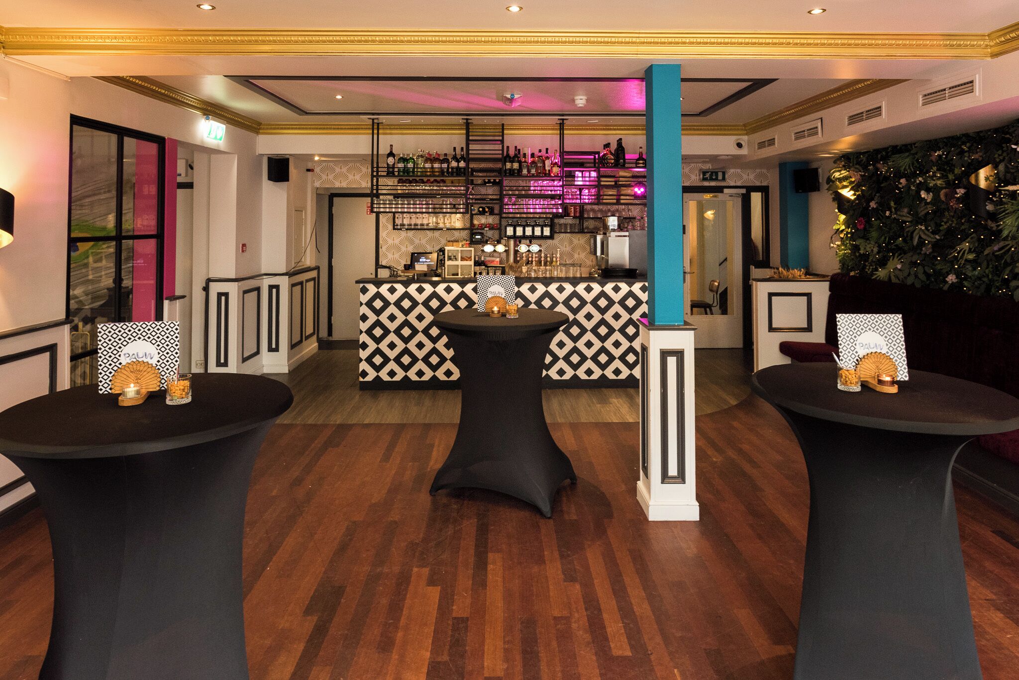 Una zona de bar con pared decorada, dos mesas redondas con centros de mesa y una barra con estantes llenos de botellas.