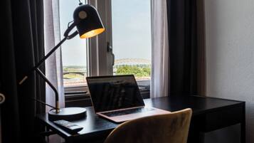 A desk with a laptop, a lamp, and a chair in a room with a view of a bridge and greenery.