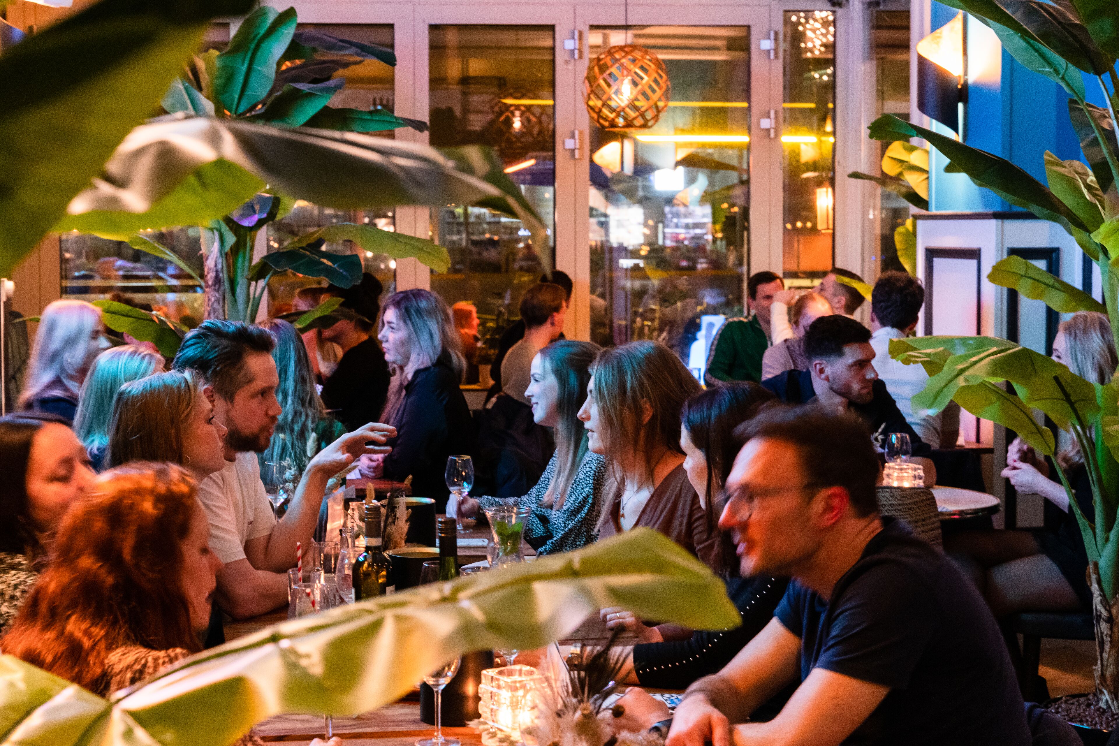 Eine Gruppe von Menschen speist in einem Restaurant mit großen Fenstern und Zimmerpflanzen.