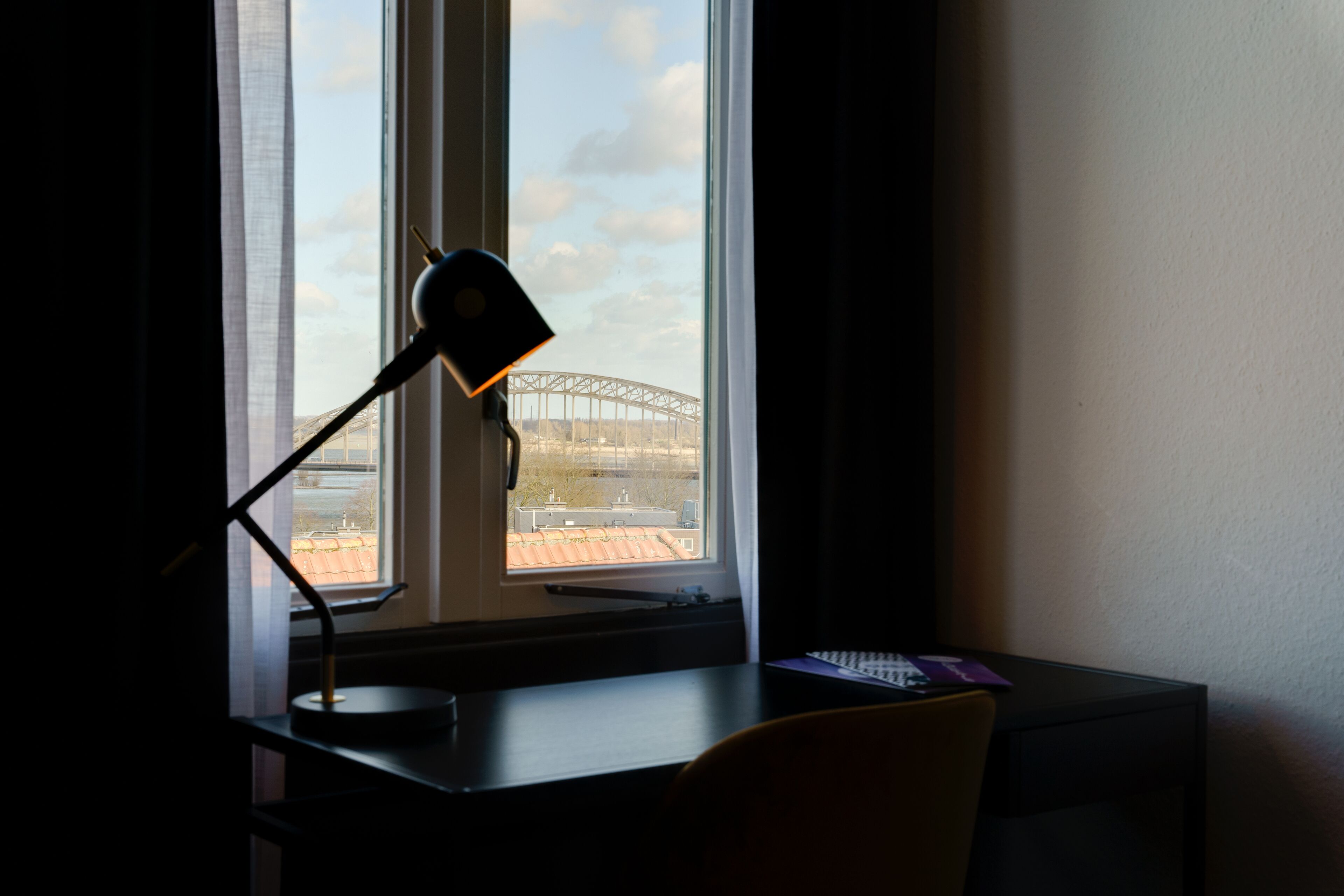 Un bureau avec une lampe, une chaise et une vue sur un pont, visible à travers une fenêtre.