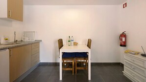 Une cuisine avec une table blanche, des chaises en bois, un meuble blanc, un extincteur et un sol carrelé.