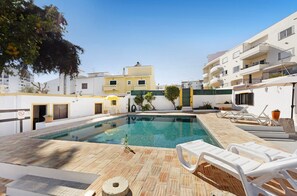 Una piscina con sillones de descanso, un suelo de baldosas y un edificio con balcones al fondo.