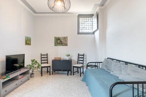 A small, well-lit room with a bed, a TV, a desk with two chairs, and artwork on the walls.