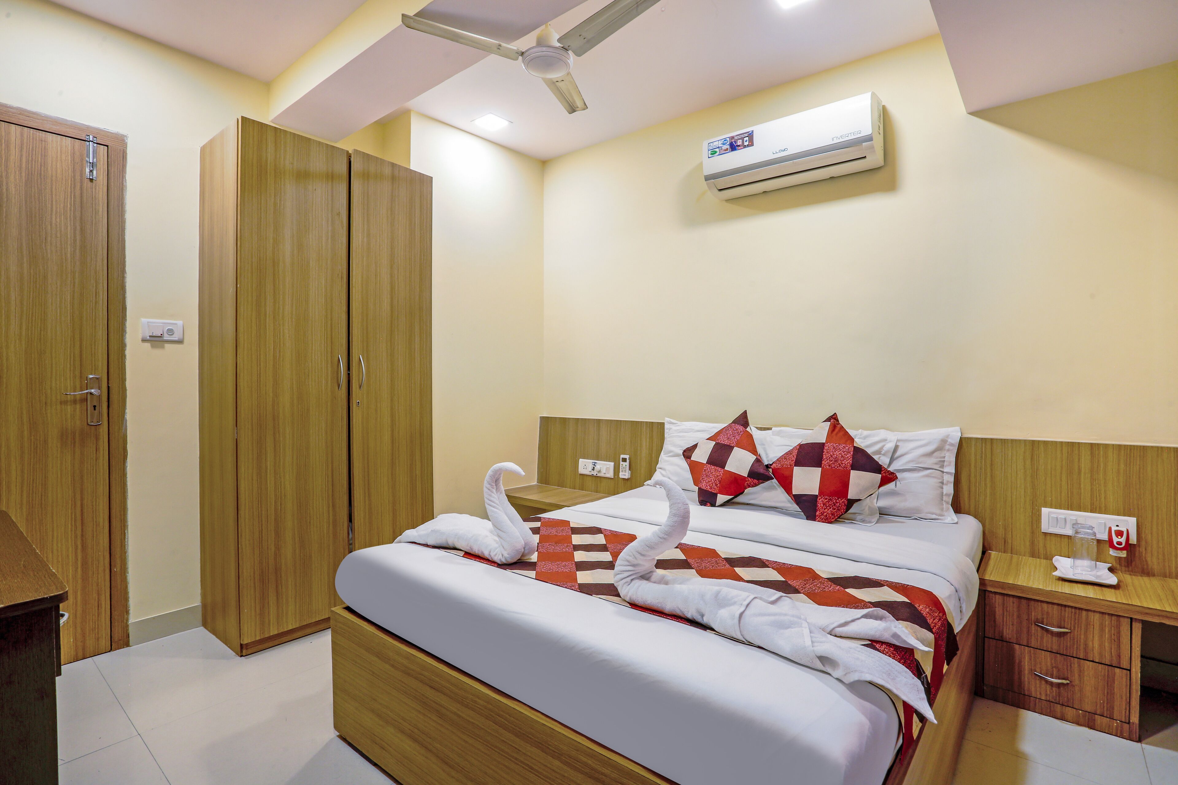A hotel room with a bed, wooden wardrobe, bedside table, and an air conditioning unit.