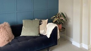 A room with a dark blue accent wall, a white ceiling with a decorative light fixture, a dark blue sofa with cushions, a small potted plant, and a white rug.
