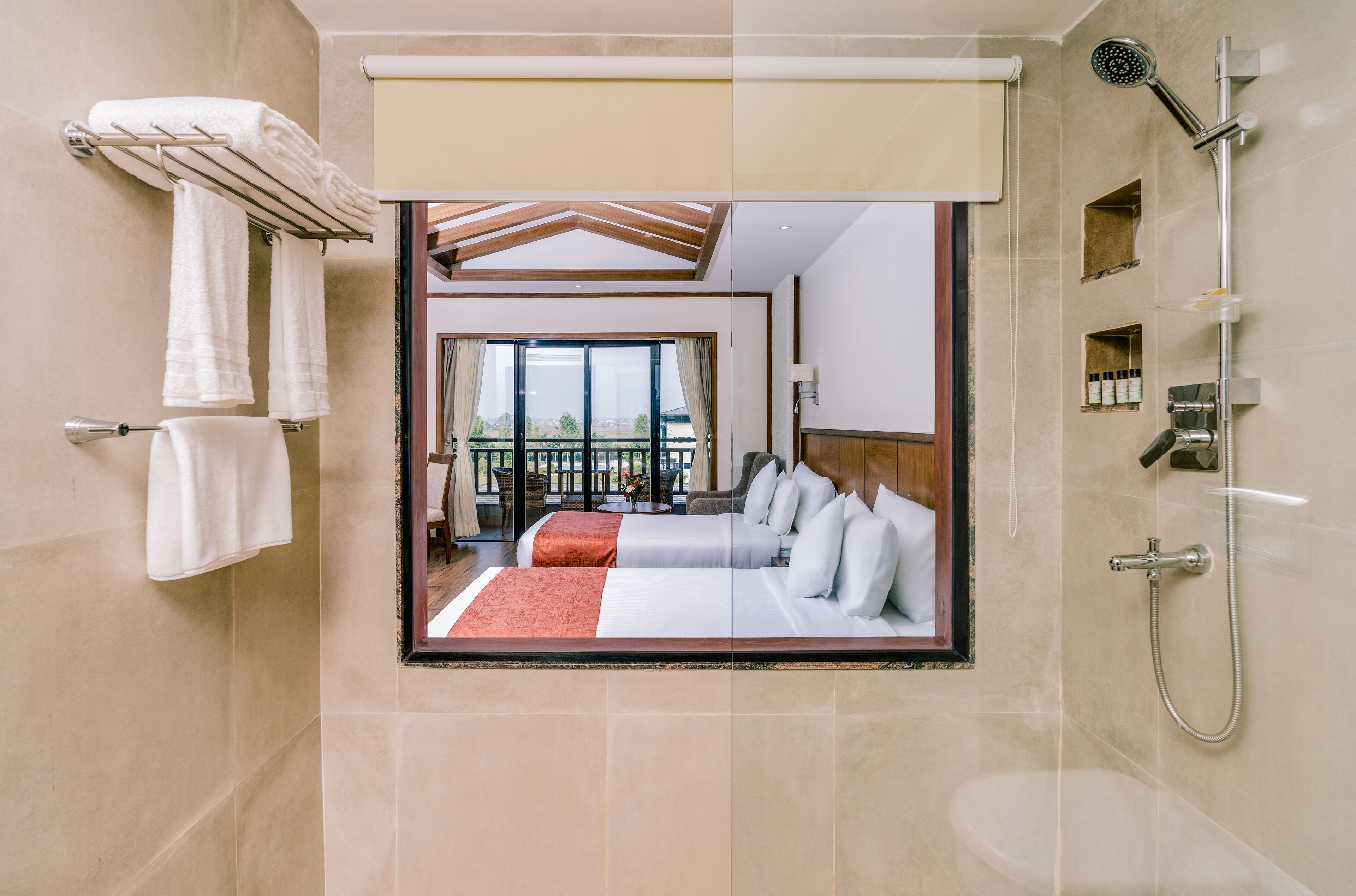 A modern bathroom with a shower, a towel rack, and a large mirror reflecting a bedroom with a bed, pillows, and a balcony view.