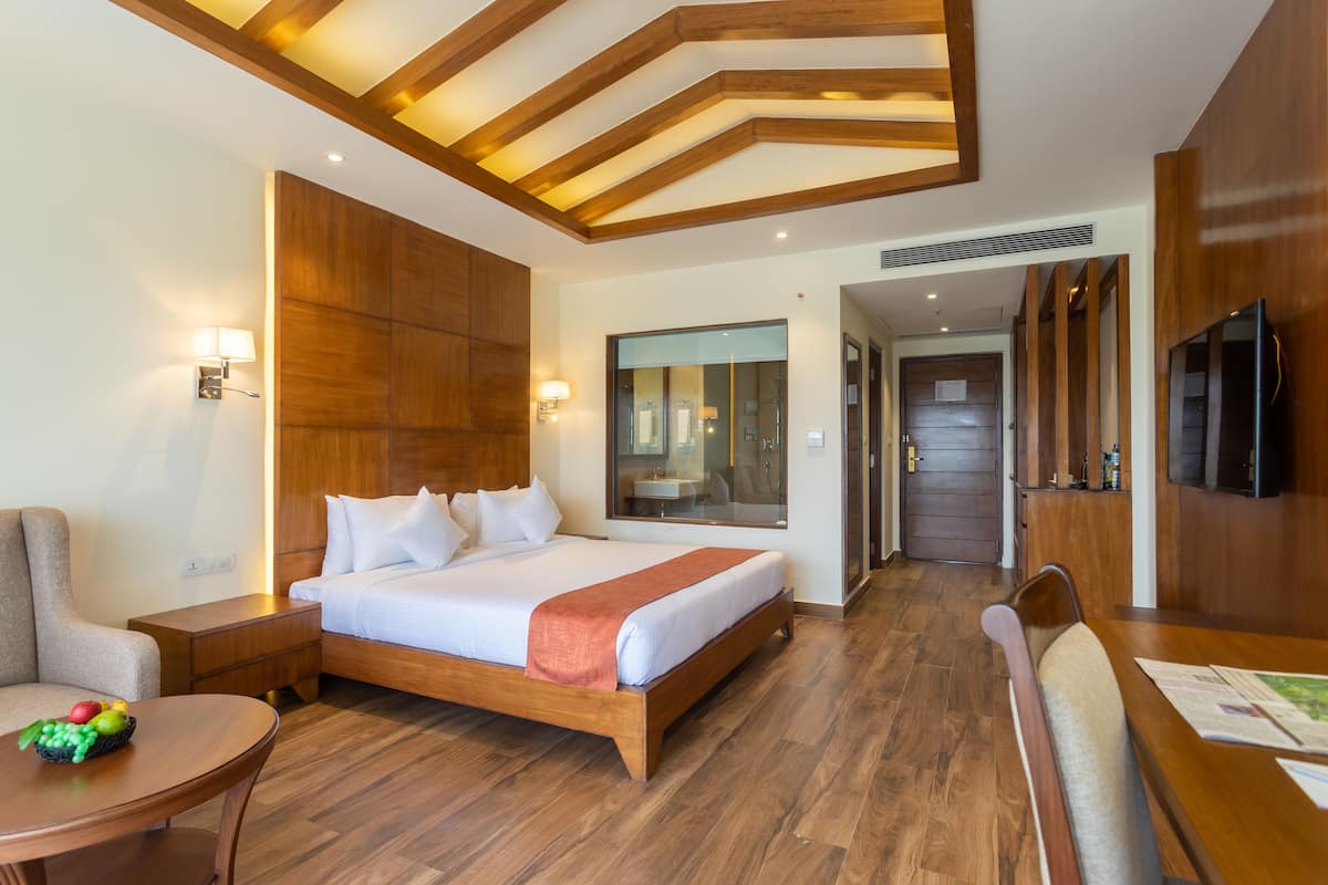 Habitación de hotel con una cama grande, cabecera de madera, mesitas de noche, televisión, mesa de comedor y vistas al baño y al pasillo.
