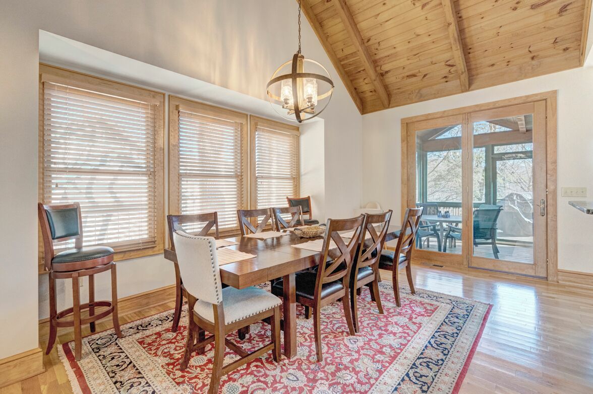 A dining area with a wooden table and chairs, large windows with blinds, a chandelier, and a view through glass doors.