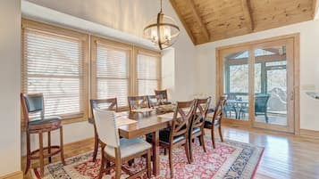 A dining area with a wooden table and chairs, large windows with blinds, a chandelier, and a view through glass doors.