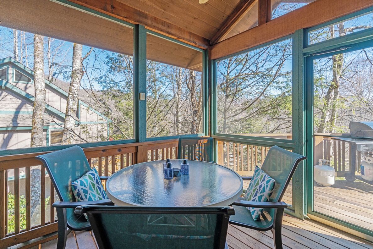 A covered outdoor seating area with a round table and chairs, large windows, and a view of trees.