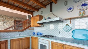 A kitchen with wooden cabinets, a gas stove, a sink, and a stone wall. - Villa Mytilene II by Wonderful Italy (Cefalù)