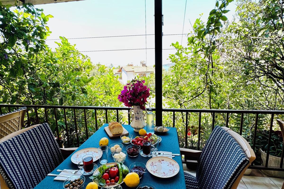 A table set for a meal with various dishes and a vase of flowers on a balcony with a view of greenery and buildings.