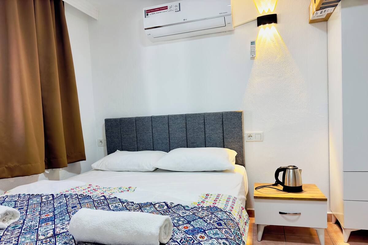 A hotel room with a bed, a nightstand with a kettle, and an air conditioning unit.