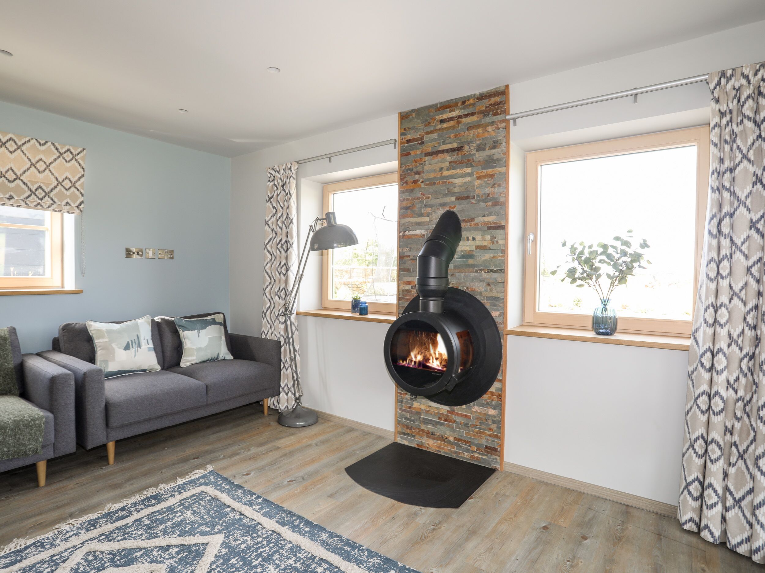 A modern living room with a fireplace, a grey sofa, a patterned rug, and large windows.