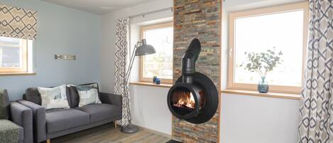A modern living room with a fireplace, a grey sofa, a patterned rug, and large windows.
