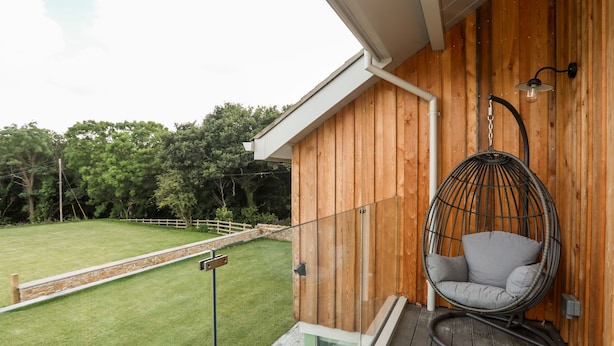 A wooden balcony with a hanging chair, a glass door leading to a grassy yard, and a stone wall.