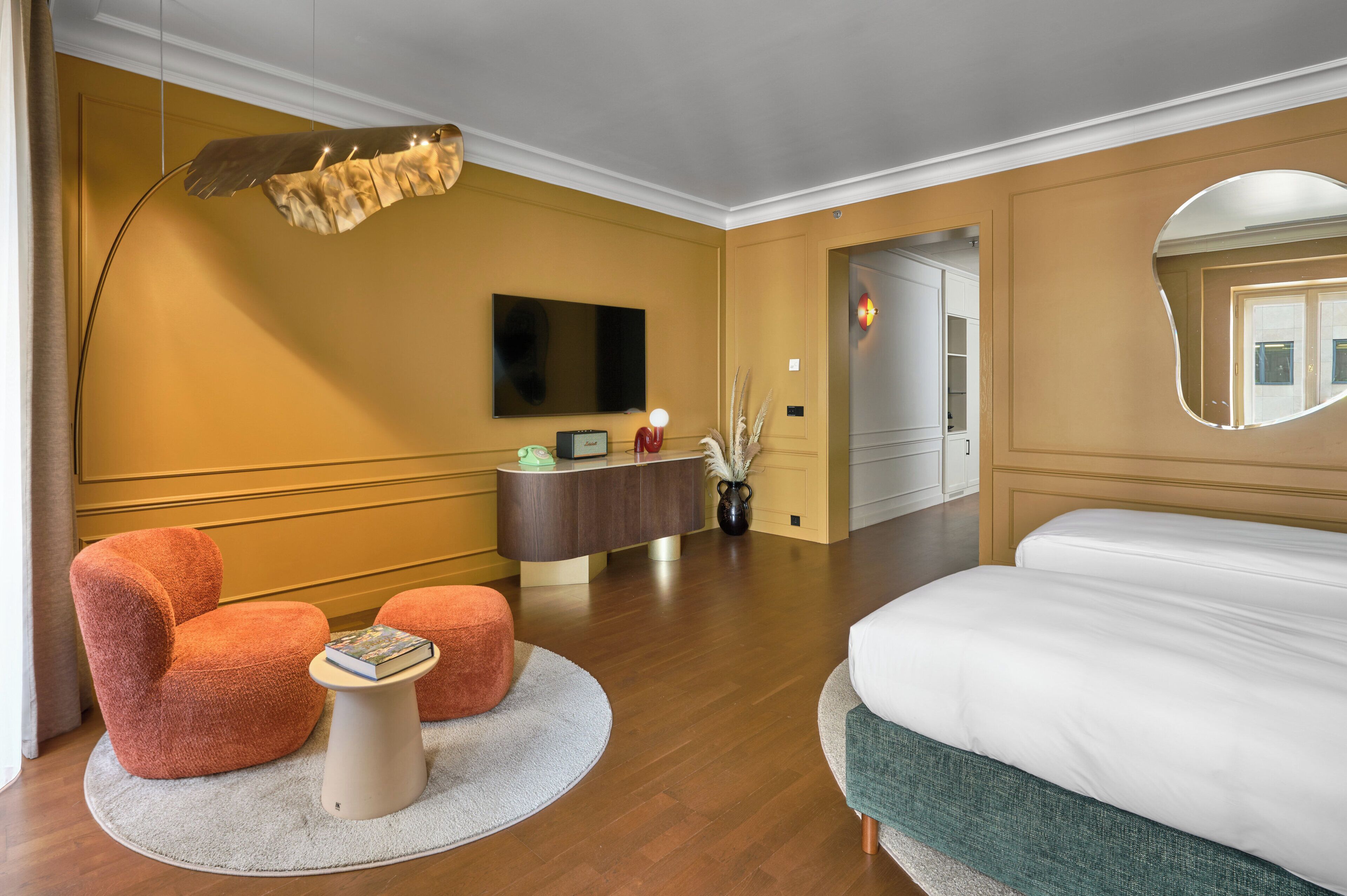 A hotel room with a large bed, a round coffee table, two orange armchairs, a wooden dresser, a flat-screen TV, and a large mirror.