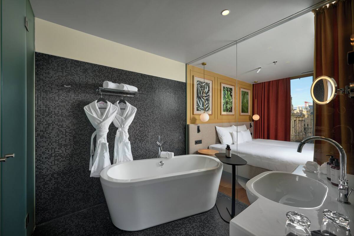 Ett modernt hotellrum med badkar, en dubbelsäng, ett sängbord, en stor spegel och utsikt över staden.