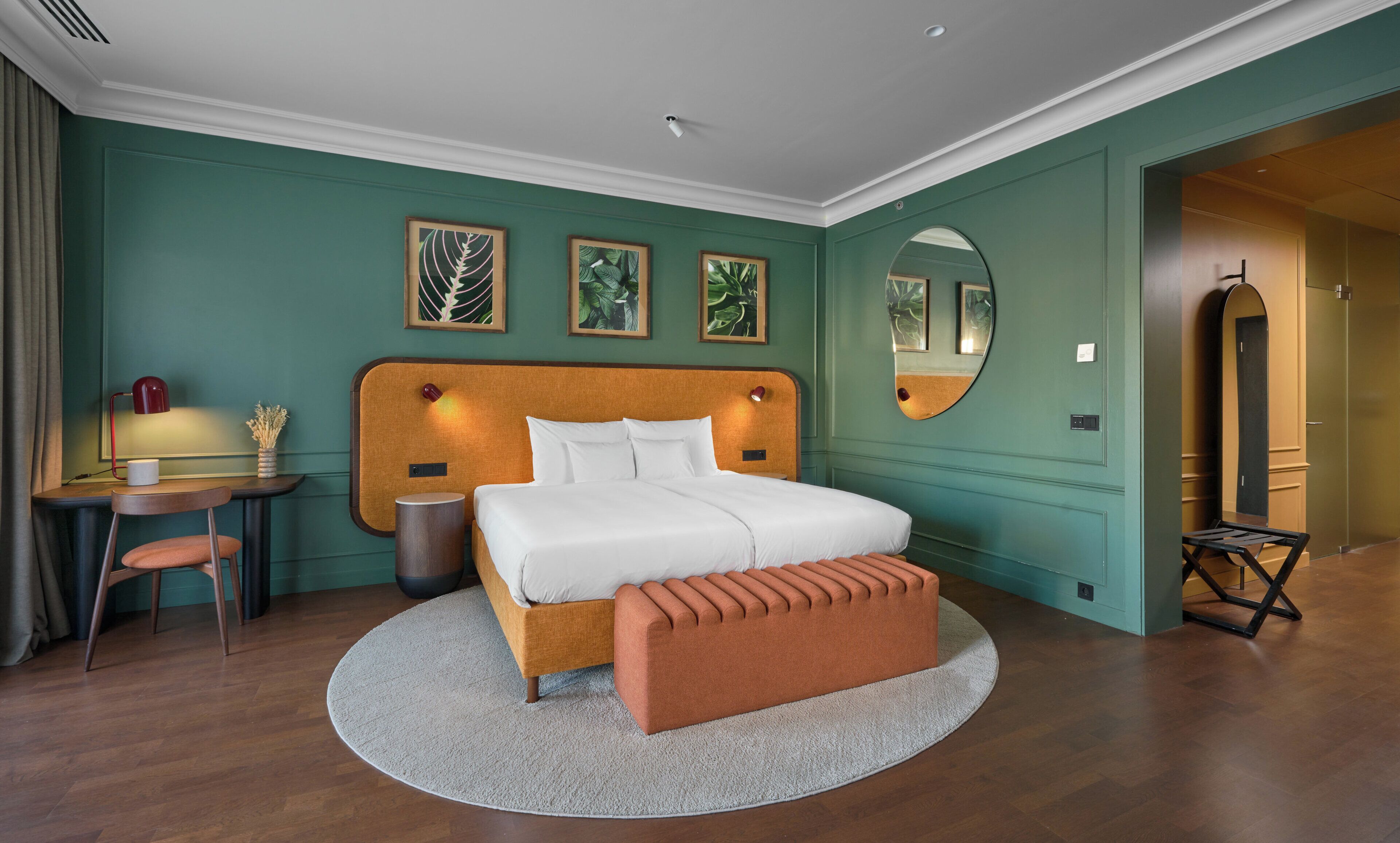 Une chambre d’hôtel avec un lit, un miroir rond, un bureau avec une lampe et une chaise.