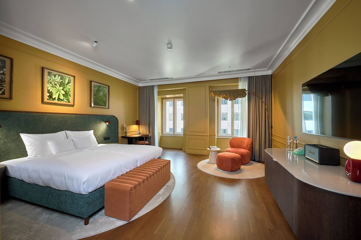 A hotel room with a large bed, a seating area, a TV, and a desk with a lamp.