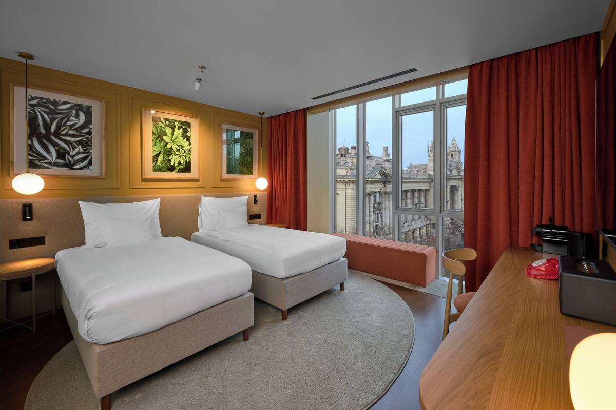 A hotel room with two beds, a desk with a computer, a view of the city, and artwork on the walls.