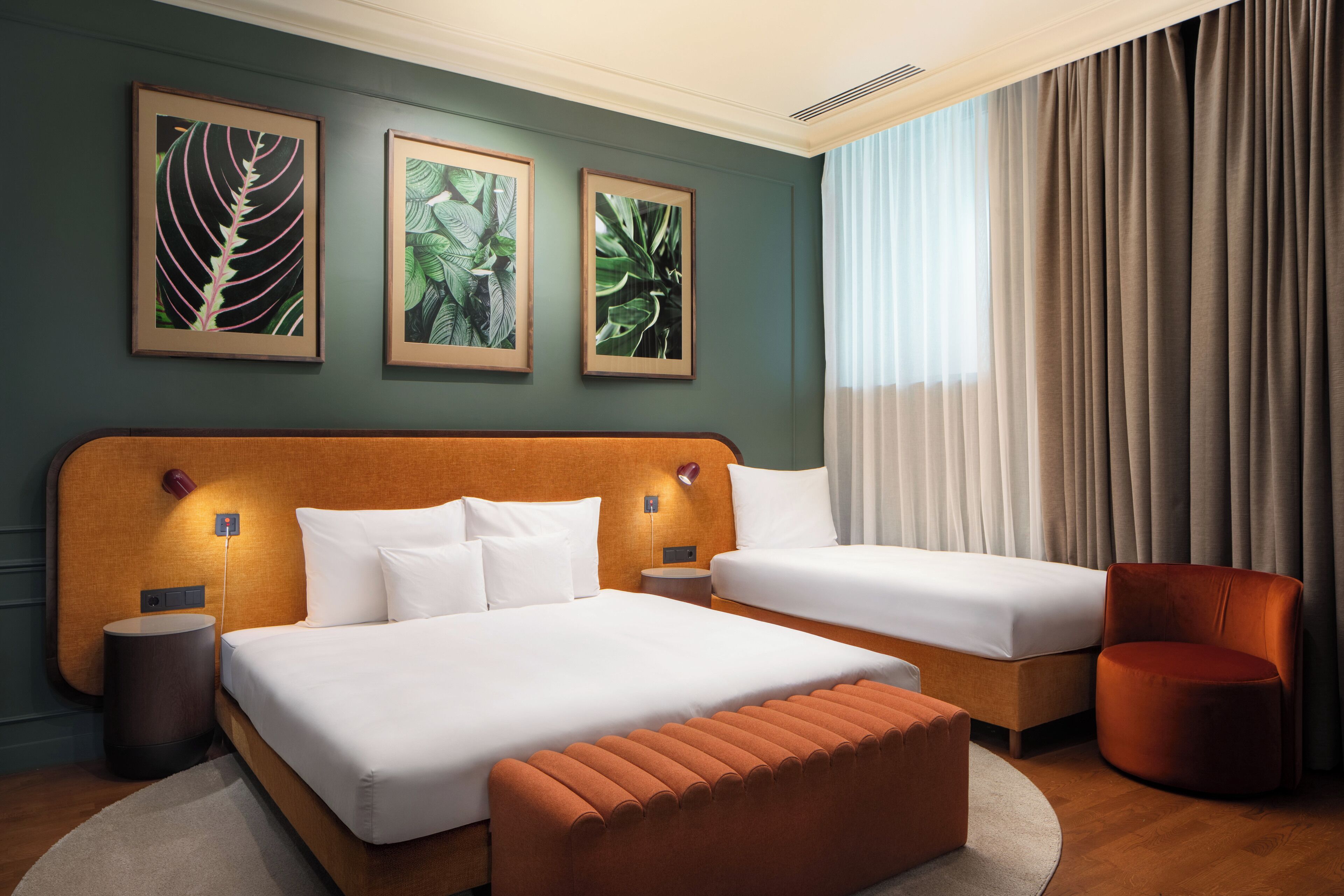 A hotel room with a large bed, a smaller bed, a round orange ottoman, and a chair. There are three framed botanical prints on the wall and a window with curtains.