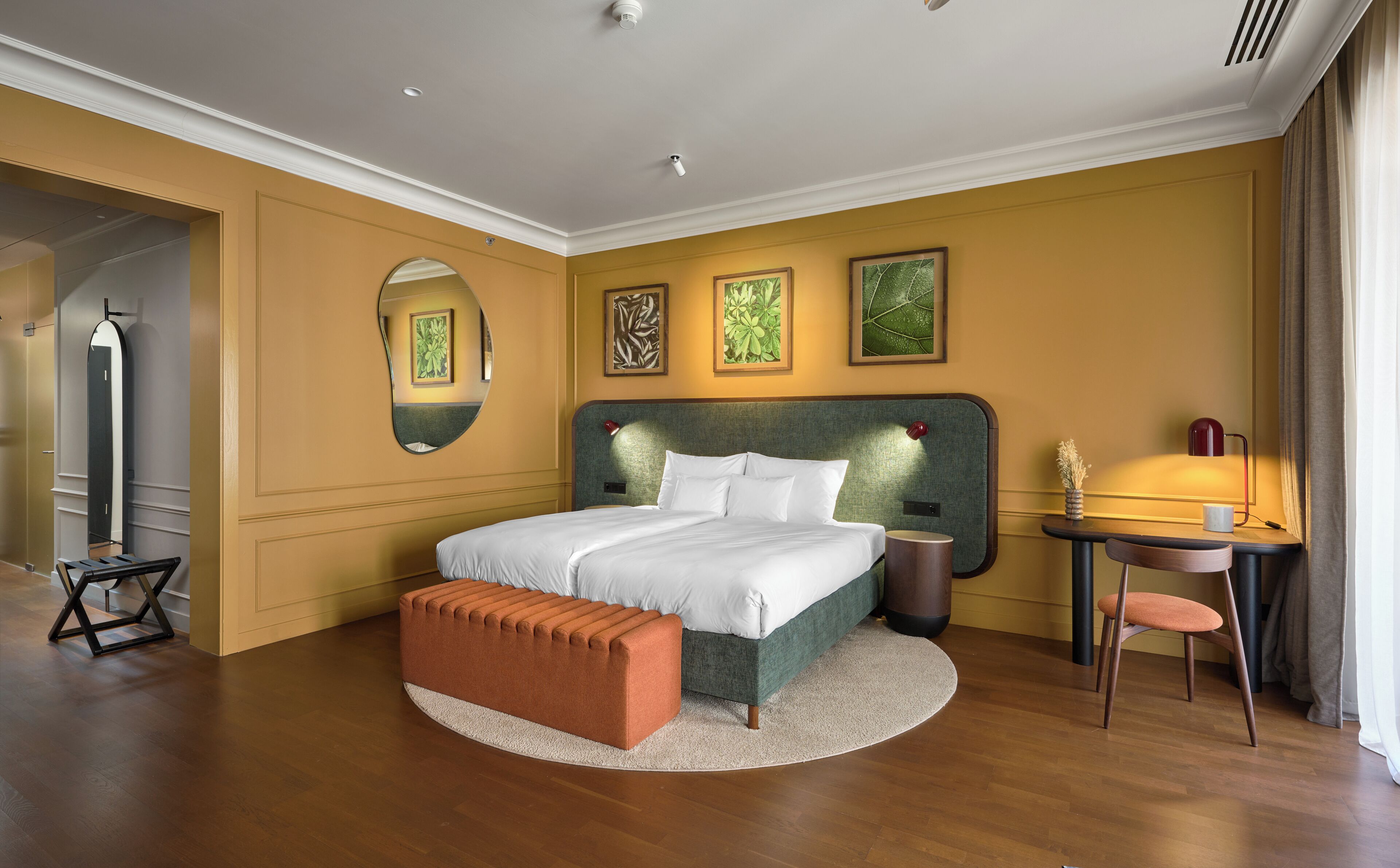 A hotel room with a bed, a round table with a chair, a small desk, a mirror, and artwork on the walls.