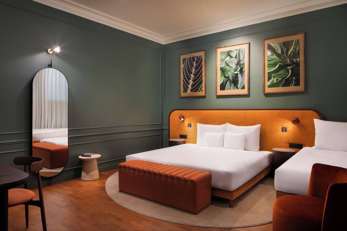 A hotel room with a large bed, two bedside tables, a round ottoman, a chair, a small side table, and framed botanical artwork on the wall.