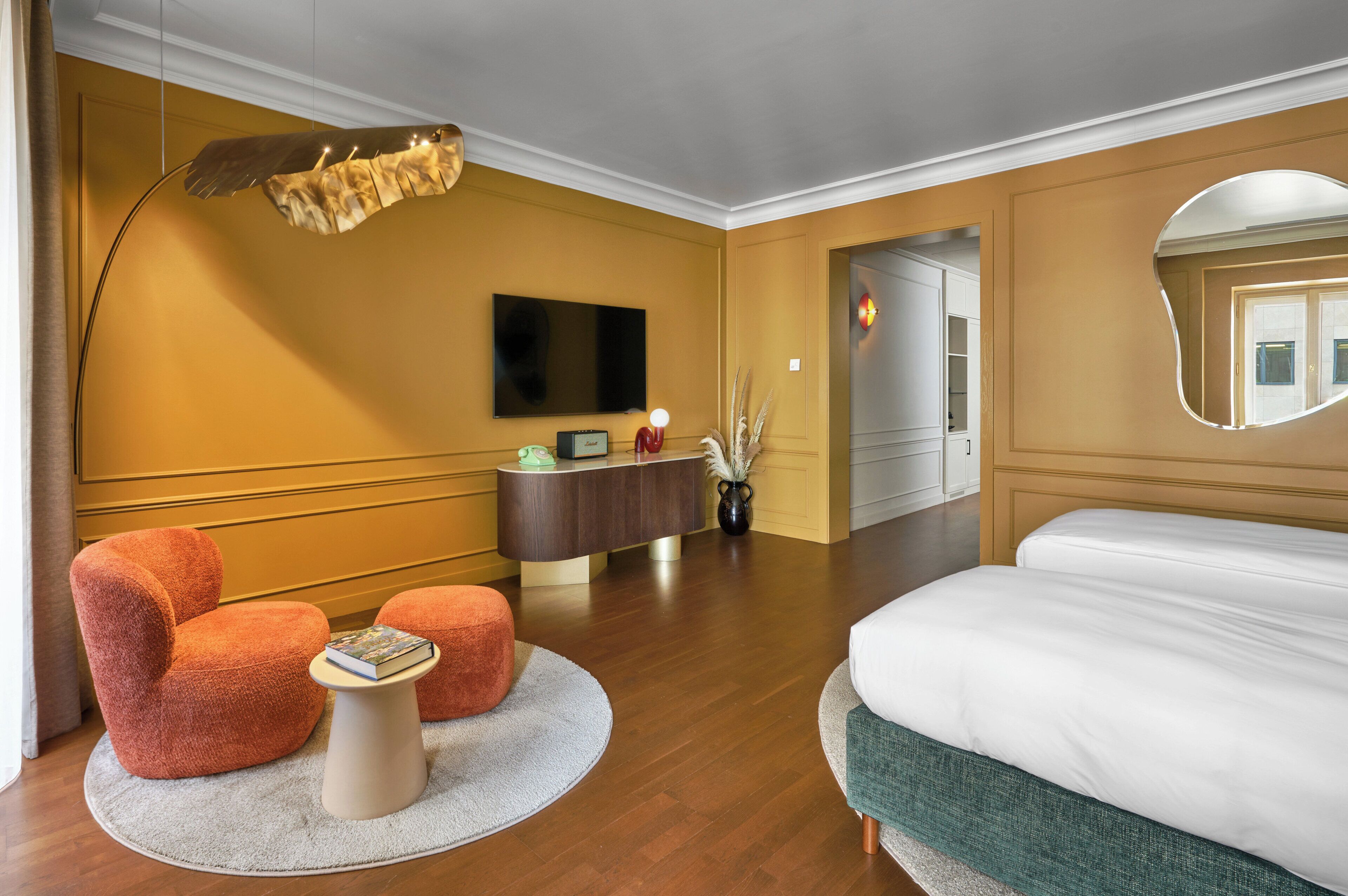 A hotel room with a bed, a desk with a flat-screen TV, two orange armchairs, a small round table, a mirror, and a decorative plant.