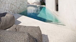 A pool area with a stone wall, a palm tree, and a white building with a window. - ANOME Mykonos (Mykonos)