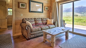 Une pièce chaleureuse avec un plafond en bois, un canapé en cuir agrémenté de coussins à motifs, une table basse en bois et une grande fenêtre donnant sur un paysage verdoyant et des montagnes.