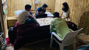 A group of people playing a board game in a room with bamboo walls and a bed.