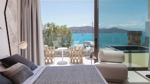 Une chambre moderne avec un grand lit, une porte vitrée donnant sur un balcon avec vue sur la mer et les montagnes.