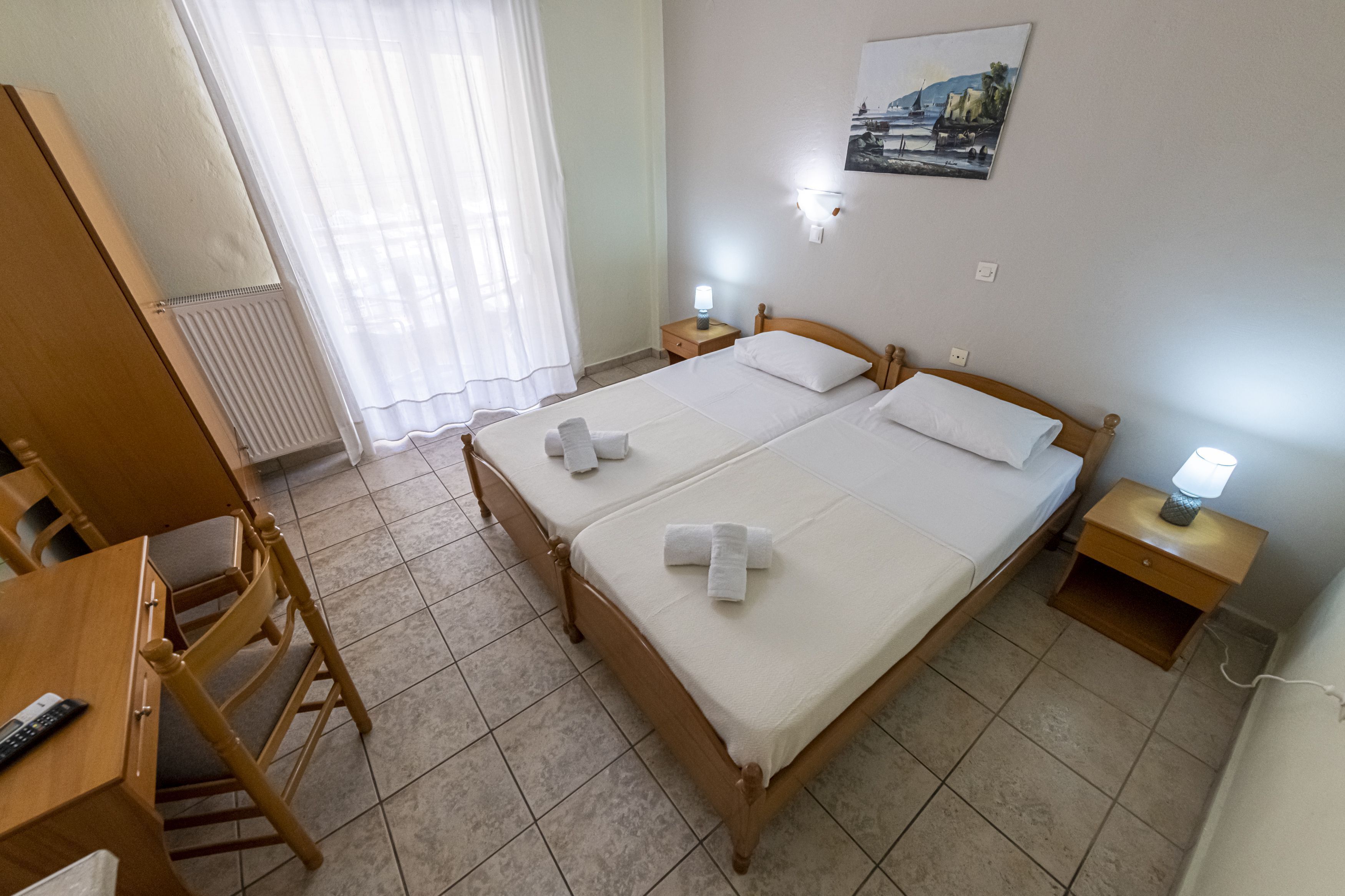 Un lit double avec du linge de lit et des serviettes blanches, une table de chevet en bois avec une lampe, un bureau avec une chaise et un radiateur.