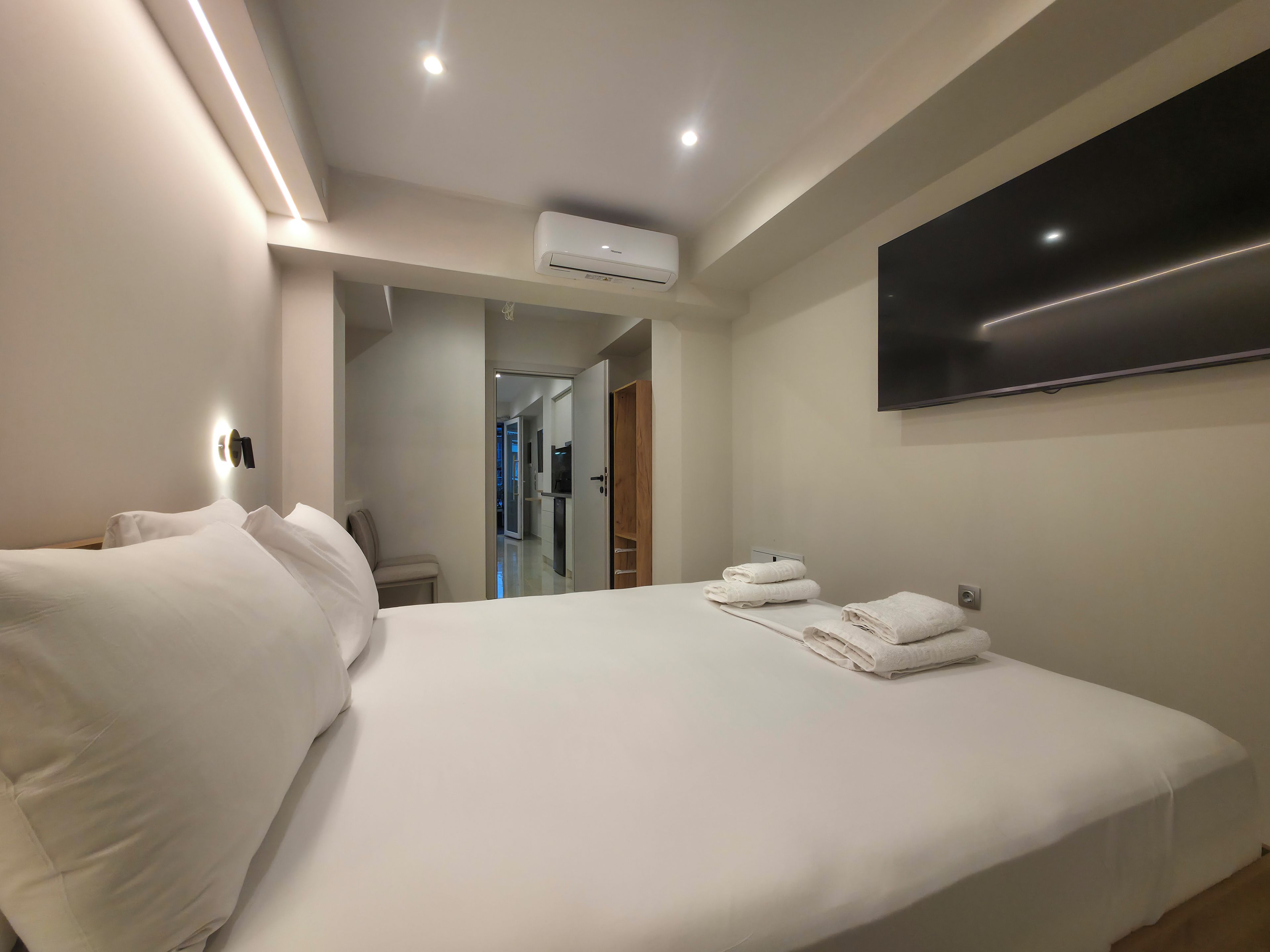 A hotel room with a bed, a flat-screen TV mounted on the wall, and a hallway leading to other rooms.