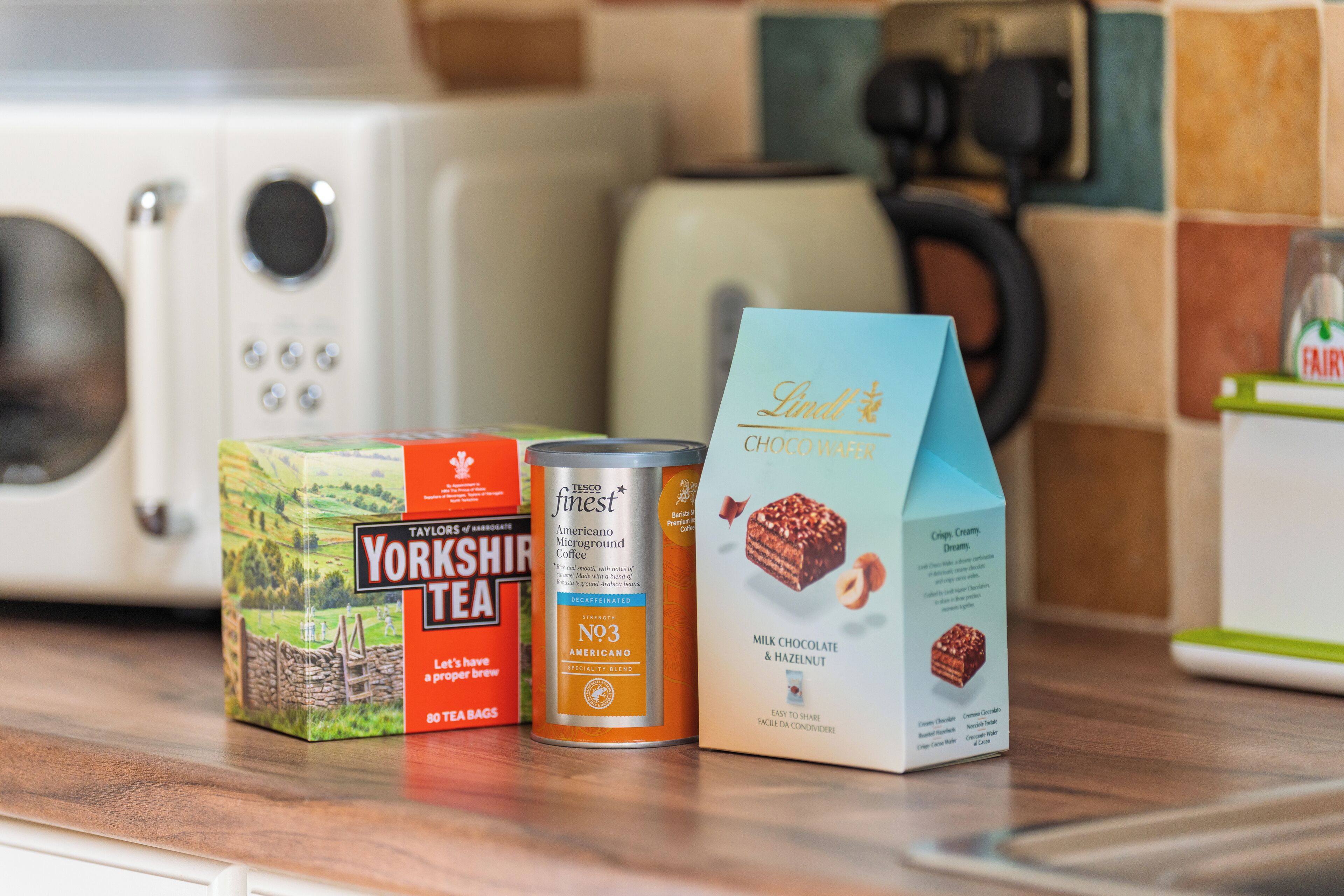 Una encimera de cocina con una caja de té Yorkshire, un bote de café Tesco Finest N93 y un cartón de chocolate con leche y avellanas de Lidl.