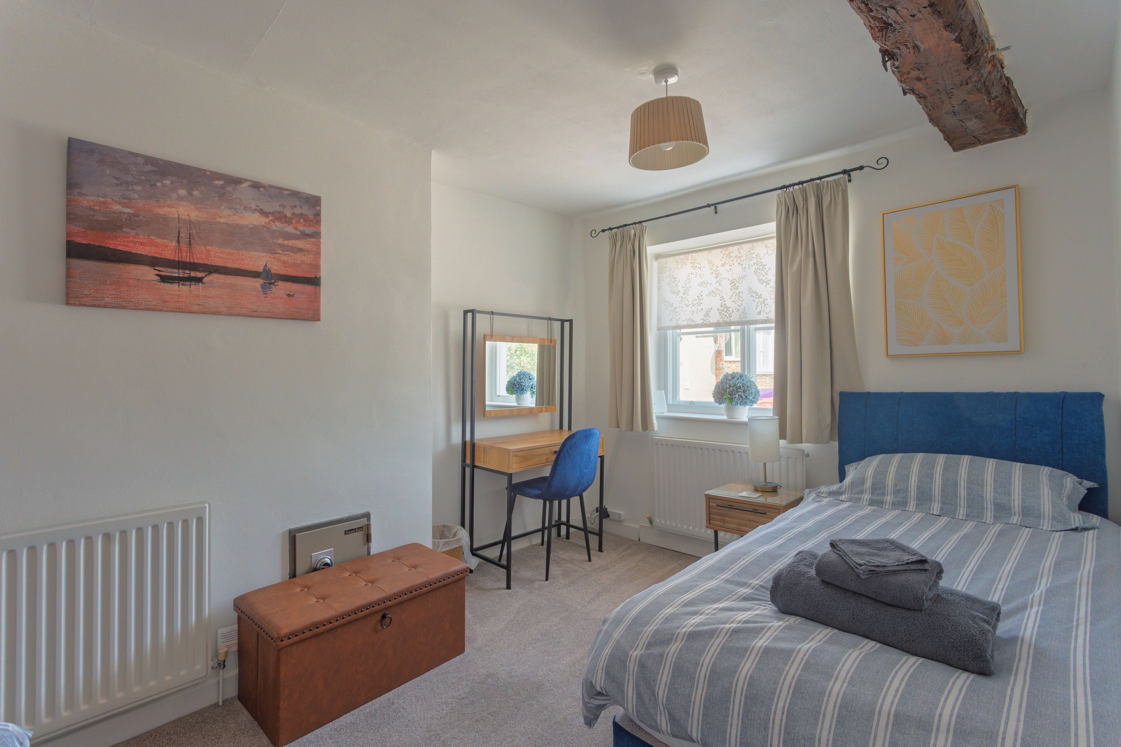 Een slaapkamer met een bed, bureau, stoel, radiator, raam met gordijnen en een schilderij aan de muur.