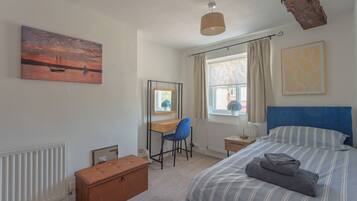 Een slaapkamer met een bed, bureau, stoel, radiator, raam met gordijnen en een schilderij aan de muur.
