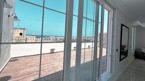 A room with large windows offering a view of a coastal area with a stone wall and a clear blue sky. - Work & Travel - Valletta Apartments (Valletta)