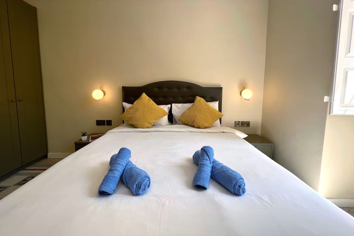 A neatly made bed with two rolled towels, a dark headboard, and two wall-mounted light fixtures.