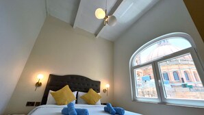 A hotel room with a bed, two wall-mounted lamps, a ceiling fan, and a large arched window with a view of a building. - Work & Travel - Valletta Apartments (Valletta)