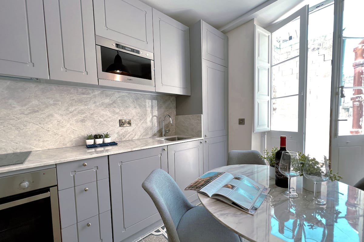 A modern kitchen with a built-in oven, a microwave, and a sink. There is a dining table with chairs and a magazine.
