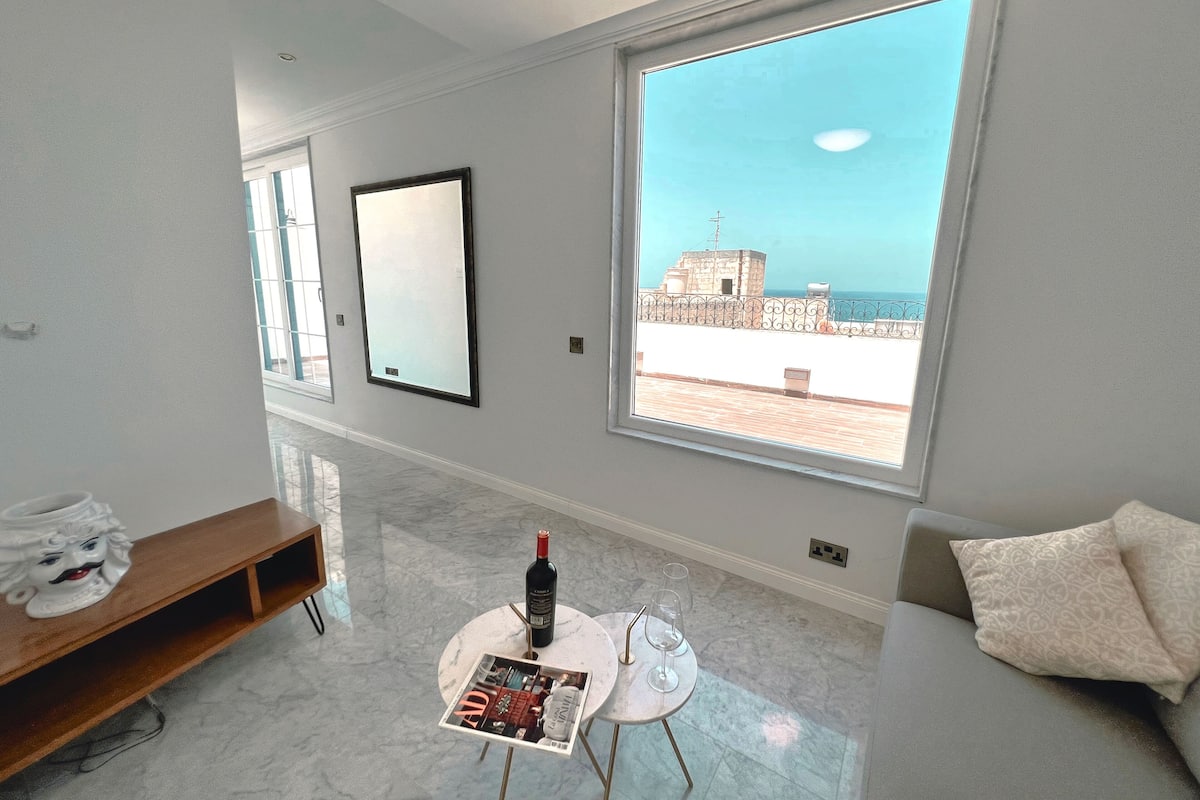 A modern living room with a large window, a marble floor, a round table with a magazine, a bottle of wine, and a sofa with a patterned cushion.