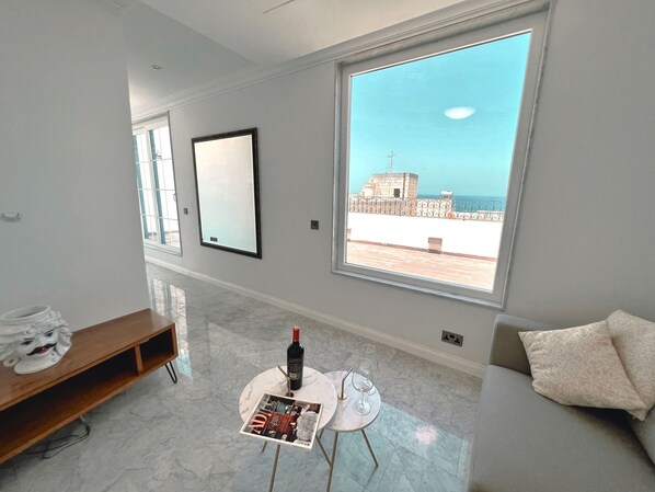 A modern living room with a large window, a marble floor, a round table with a magazine, a bottle of wine, and a sofa with a patterned cushion.