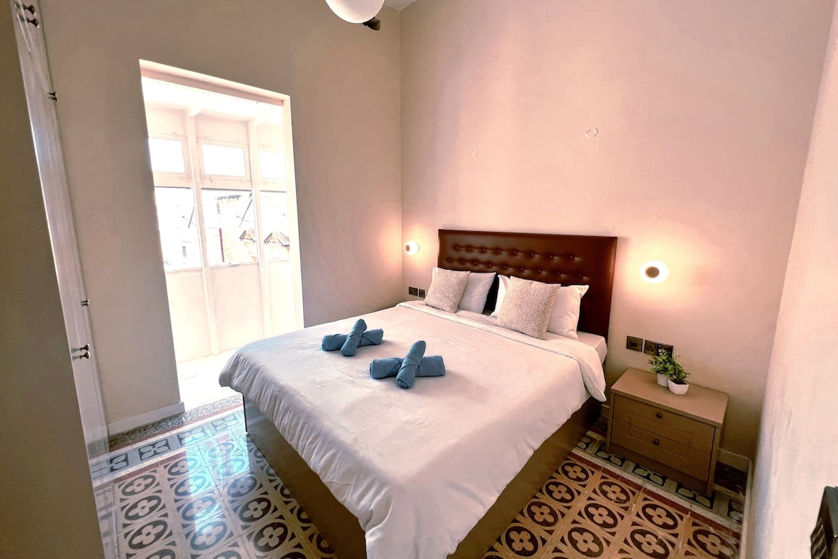 A neatly made bed with a headboard, a bedside table with a lamp, and a patterned floor.