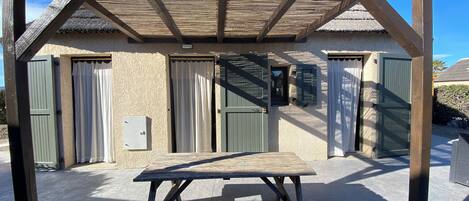 A wooden pergola with a table in front, a building with green shutters, and a clear sky.