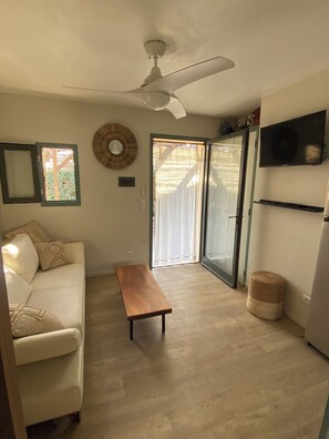 A living room with a white sofa, a wooden bench, a ceiling fan, a TV mounted on the wall, and a sliding glass door leading to a balcony. - Fisherman's Island Mas (Le Barcarès)