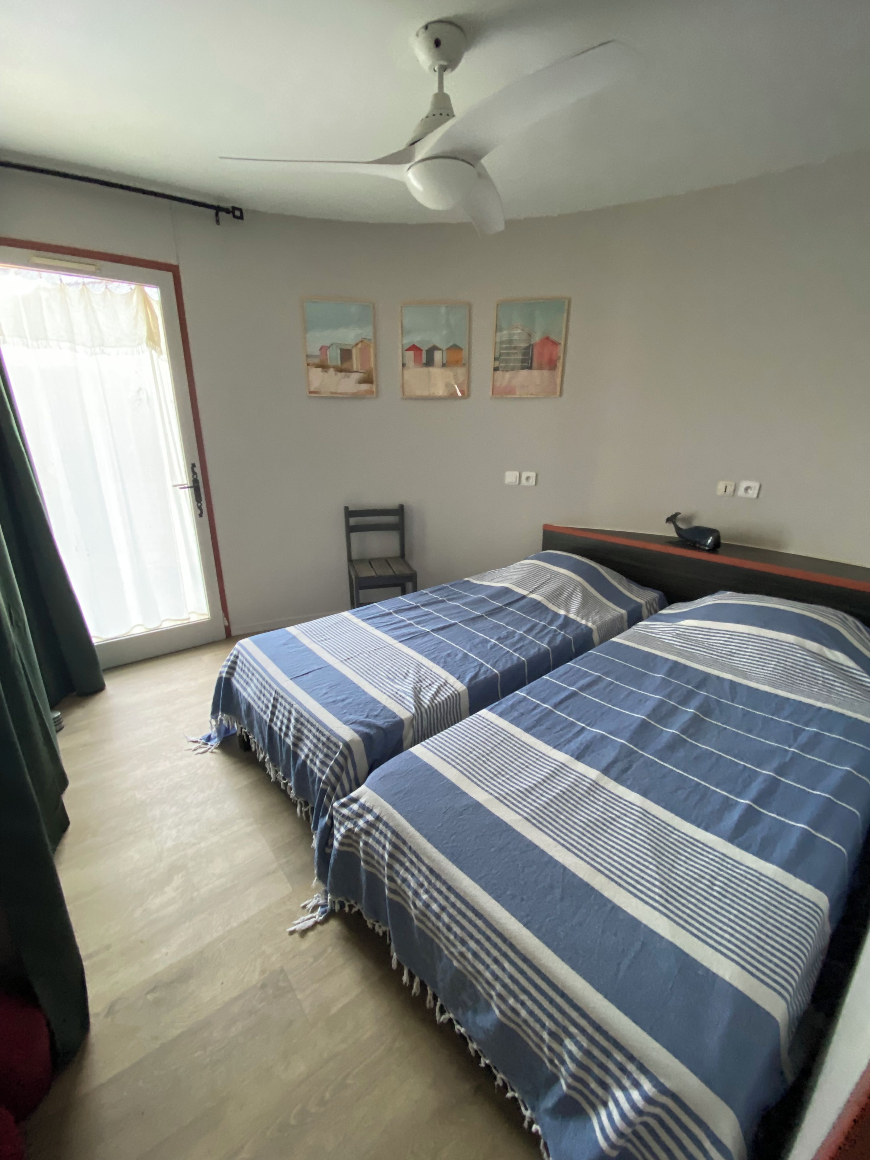 A bedroom with two beds, a ceiling fan, a window with curtains, and three wall-mounted paintings.